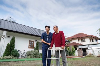 İlgili genç bir bakıcı yaşlı bir adama yürüteçle yardım eder, çağdaş bir evin yanındaki manzaralı bahçede hareket ve kendine güven sağlar..