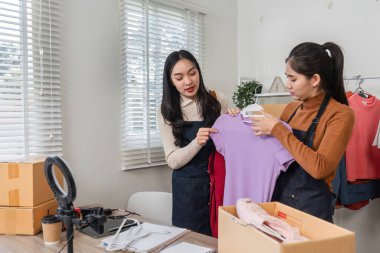 İki kadın canlı mor bir bluz ve kırmızı bir giysi sunarken dostça bir tartışma içindeler. Parlak bir ev ofisinde çevrimiçi dükkanlarına hazırlanıyorlar..