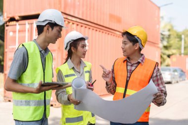 Aralarında erkekler ve bir kadın denetçinin de yer aldığı çeşitli inşaat işçileri ekibi planları inceleyip konteynırlarla çevrili bir inşaat sahasında fikirlerini paylaşıyor..