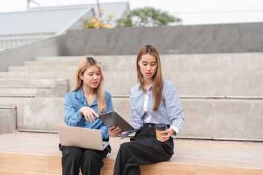 Bir tablet ve dizüstü bilgisayar üzerinde işbirliği yapan iki farklı kadın, takım çalışmasını ve üretkenliği vurgulayarak, açık hava çalışma alanında kahvenin tadını çıkarıyorlar..