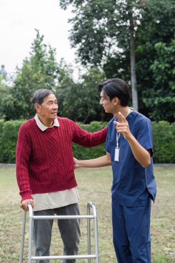 Şefkatli genç bir bakıcı yaşlı bir adamla yürüteç kullanarak ilişki kurar, yemyeşil bir bahçe ortamında anlamlı bir konuşma yapar, bağlantıyı güçlendirir ve duygusal destek sağlar..