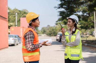 İki inşaat işçisi, bir erkek işçi ve bir kadın yönetici, inşaat alanında güvenlik yelekleri ve başlıklar giyerken proje detayları hakkında iletişim kuruyorlar..