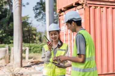 İki inşaat mühendisi, nakliye konteynırları yakınlarındaki planları incelerken, takım çalışmasını ve koordinasyonunu vurgularken, proje güncellemelerini iletmek için radyo kullanıyor..