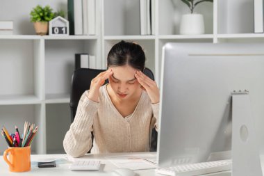 Bilgisayar masasında çalışırken stres ve baş ağrısı çeken bir kadın modern bir çalışma alanında ofis sendromunun zorluklarını yansıtıyor..