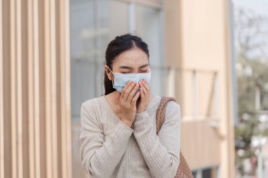 Koruyucu maske takan bir kadın, çağdaş bir şehir ortamında dışarıda dikilirken kaygılarını dile getiriyor..
