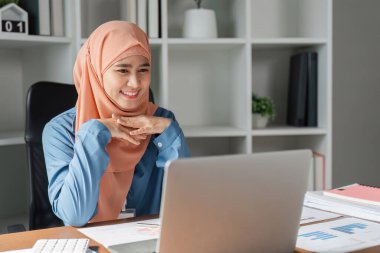 Başörtülü Müslüman bir kadın laptopuyla mutlu bir şekilde meşgul oluyor ve çağdaş ofis çalışma alanındaki belgeleri inceliyor..