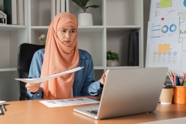 Modern ofis ortamında bilgisayarındaki finansal belgeleri incelerken endişeli görünen tesettürlü çeşitli Müslüman bir kadın..