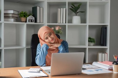 Tesettürlü, tesettürlü, neşeli bir Müslüman kadın laptopuyla çalışırken neşeli ve modern ofis ortamında çalışıyor..