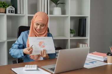 Başörtülü Müslüman bir kadın, çağdaş ofis ortamındaki bir video konuşmasında bunu açıklarken bir bütçe pasta grafiğine işaret ediyor..