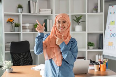 Başörtülü Müslüman bir kadın, modern ofisinde bitkiler ve iş malzemeleriyle çevrili bir şekilde gülümserken iki eliyle işaret ediyor..