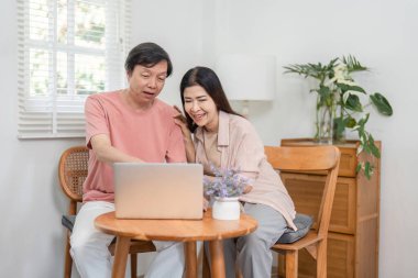 Çevrimiçi aktivitelere katılan yaşlı bir çift rahat evlerinde dizüstü bilgisayar kullanırken gülümsüyor..