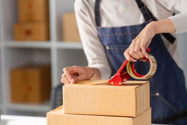 Genç bir kadının elleri, kutularla dolu, parlak ve düzenli bir çalışma alanına bantla bir nakliye kutusunu mühürlüyor..