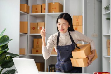 Neşeli genç bir bayan girişimci, çalışma alanındaki başarısını kutluyor. Dizüstü bilgisayarı üzerinde çalışırken elinde paketler tutuyor..
