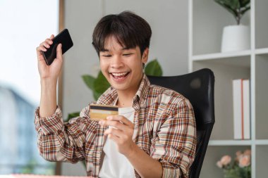 Neşeli genç bir adam akıllı telefon ve kredi kartı kullanarak başarılı bir finansal yönetimi kutluyor ve mutluluğu gösteriyor..