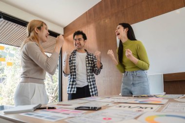 Üç kişilik neşeli bir ekip başarılarını parlak bir ofiste kutluyor, takım çalışmasını ve işbirliğini ön plana çıkarıyor..