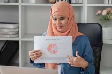 Modern bir ofis masasında oturmuş mali yönetim becerilerini sergileyen çeşitli müslüman bir kadın, bir bütçe pasta grafiği sunuyor..