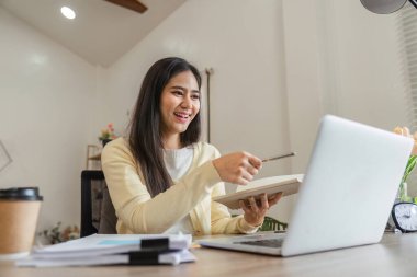 Gülümseyen Asyalı bir kadın dizüstü bilgisayarıyla uğraşırken elinde bir defter ve kalem tutuyor, üretken ofis ortamında yaratıcılık ve coşku sergiliyor..