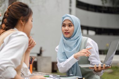 Başörtüsü takan bir kız öğrenci laptopunu açık havada bir arkadaşına gösterirken, takım çalışmasını ve akademik desteği somutlaştıran kavramları tutkuyla anlatıyor..