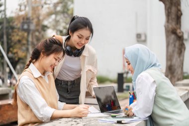 Bir grup kız öğrenci, dışarıda bir çalışma alanında dizüstü bilgisayar ve not defteriyle çalışmalar üzerinde işbirliği yaparak, takım çalışmasını ve eğitime olan bağlılığını gösteriyorlar..