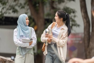 İki farklı kız öğrenci, biri tesettür takıyor, kampüste birlikte yürüyor ve gülümsüyor, kitap taşıyor, arkadaşlığı ve akademik kutlamayı sembolize ediyor.