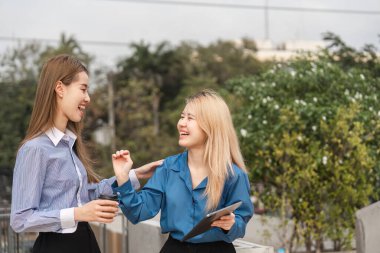 İki farklı kadın, canlı, yeşil bir ortamda ellerinde bir tablet ve kahve tutarken mutlu bir şekilde sohbet ediyorlar..