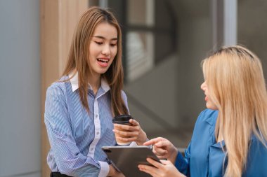 İki farklı kadın fikirleri tartışıyor, biri kahve tutuyor, diğeri tablet kullanıyor, modern çalışma ortamında..