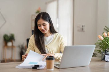 Evrakları düzenleyen, rahat bir ofiste dizüstü bilgisayarıyla çalışan, kendini işine adamış ve üretken bir iş ortamı sunan konsantre bir genç kadın..