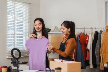 Genç bir kadın çevrimiçi kıyafet işini yönetirken renkli tişörtler sergiliyor, müşteri ilişkileri ve ürün çeşitliliğini vurguluyor.