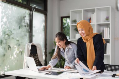 Finans sektöründe iki farklı kadın birlikte finansal raporları neşeyle gözden geçirir, işbirliği ve başarılarını vurgularlar.