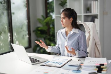 Bir video görüşmesi sırasında stratejileri tartışırken, parlak bir ofiste iletişim ve işbirliğini vurgulayan bir finans profesyoneli jestler yapar.