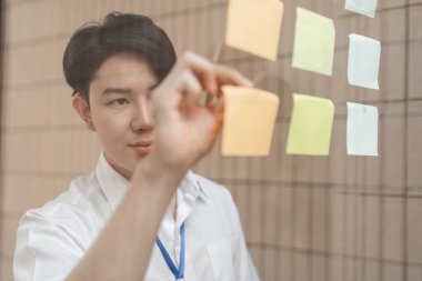 Genç bir profesyonel aktif olarak cam bir duvara renkli yapışkan notlar yerleştiriyor, fikirleri düzenliyor ve parlak ofis alanındaki iş akışını artırıyor..