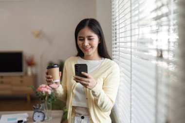 Genç bir kadın elinde kahve fincanıyla akıllı telefonunu kullanarak gülümsüyor. Parlak ve davetkar bir ofiste vakit geçiriyor..
