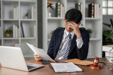 Genç bir Asyalı avukat masasında stresli bir şekilde duruyor, yasal belgeleri ve dava dosyalarını incelerken aynı zamanda avukatlık bürosundaki zorlukları yönetiyor..