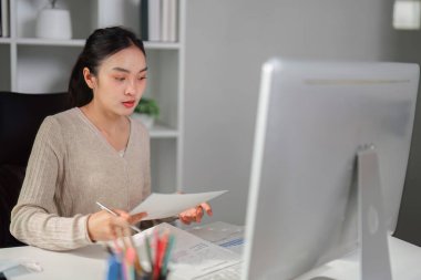 Genç bir kadın masasında otururken evrakları inceliyor, modern ofis ortamında konsantrasyon ve profesyonellik gösteriyor..