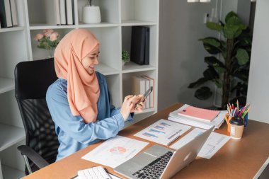 Modern ofis ortamında mali belgelerle dolu bir masada otururken akıllı telefon kullanan tesettürlü bir Müslüman kadın..