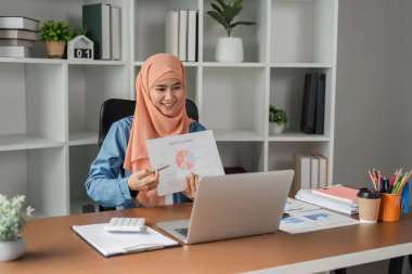 Başörtüsü takan Müslüman bir kadın, şık bir ofis ortamında video görüşmesi yaparken turta grafiği içeren bir rapor sunar..