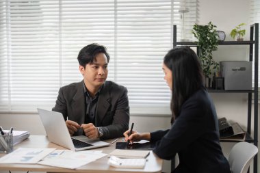 Bir erkek ve kadın profesyonel, iyi aydınlatılmış bir ofiste dizüstü bilgisayar ve evrak işleri ile finansal stratejiler hakkında bir tartışmaya girişirler..