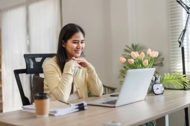 Çiçekler ve yeşilliklerle çevrili güneşli bir ofiste dizüstü bilgisayarıyla meşgul gülümseyen genç bir kadın pozitiflik ve verimlilik yayıyor..