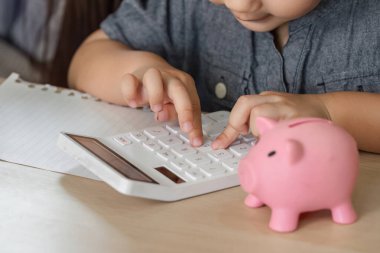 Bir çocuk, bir kumbaranın yanındaki hesap makinesiyle iletişim kurar. Finansal okur-yazarlığı ve tasarruf yapmanın önemini destekler..