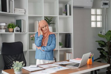 Başörtülü Müslüman bir kadın, raporlarını ve çizelgelerini çağdaş ofis ortamında dizüstü bilgisayarla incelerken stresli görünüyor..