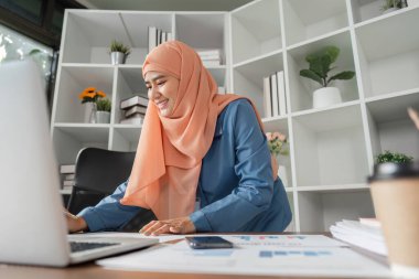 Tesettürlü, gülümseyen Müslüman bir kadın masasında bilgisayarla çalışırken finansal belgeleri düzenliyor..