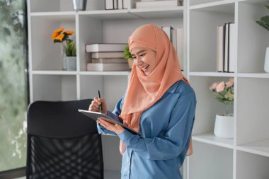 Tablet kullanan tesettürlü gülümseyen Müslüman bir kadın modern ofisinde bitkilerle ve organize raflarla çevrili notlar alıyor..