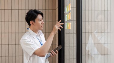 Genç bir profesyonel, elinde bir akıllı telefon tutarken cam duvardaki renkli yapışkan notları analiz ederek görevleri organize etmekle meşgul.