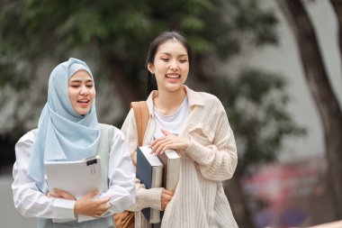 İki farklı üniversite öğrencisi, biri tesettür içinde, kampüste kitap taşırken eğleniyorlar ve arkadaşlıklarını paylaşıyorlar..
