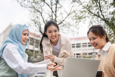 Üç çok kültürlü kız öğrenci, açık havada canlı bir akademik ortamda fikirlerini paylaşırken bir dizüstü bilgisayarda gülümsüyor ve işbirliği yapıyorlar..