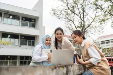 Üç çok kültürlü kız öğrenci dışarıda bir laptopta proje detaylarını gözden geçirip, kampüste takım çalışması ve işbirliği sergilediler..