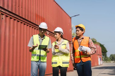 Çeşitli inşaat ekibi erkek ve kadın çalışanları, bir tablet ve nakliye konteynırlarıyla çevrili bir iş sahasının planlarını incelerken proje özelliklerini tartışıyor..