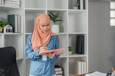 Başörtülü Müslüman bir kadın, çağdaş bir ofis ortamında dururken iş belgelerinden notlar yazıyor..