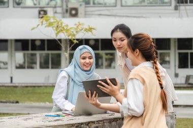 Üç farklı kız öğrenci, ortak öğrenme ortamında bir dizüstü bilgisayar ve tablet kullanarak açık havada bir proje üzerinde tartışıyorlar..