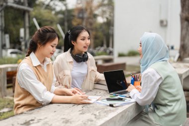 Bir grup çok kültürlü kadın üniversite ortamında dışarıda otururken fikirlerini dizüstü bilgisayar ve defterlerle paylaşıyorlar..